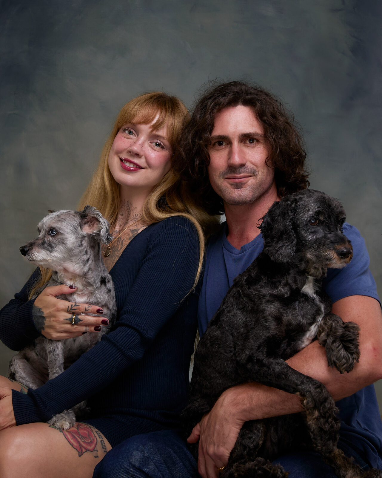 A woman and a man seated together on a dark painterly studio backdrop, each holding one of their dogs: a grey scruffy terrier and a black curly-coated dog